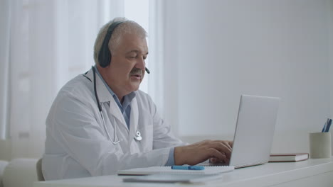 Der-Männliche-Arzt-Spricht-Mit-Dem-Patienten-Online-Per-Videoanruf-Auf-Dem-Laptop,-Hört-über-Kopfhörer-Zu-Und-Füllt-Den-Medizinischen-Bericht-Aus-Der-Ferne-Aus